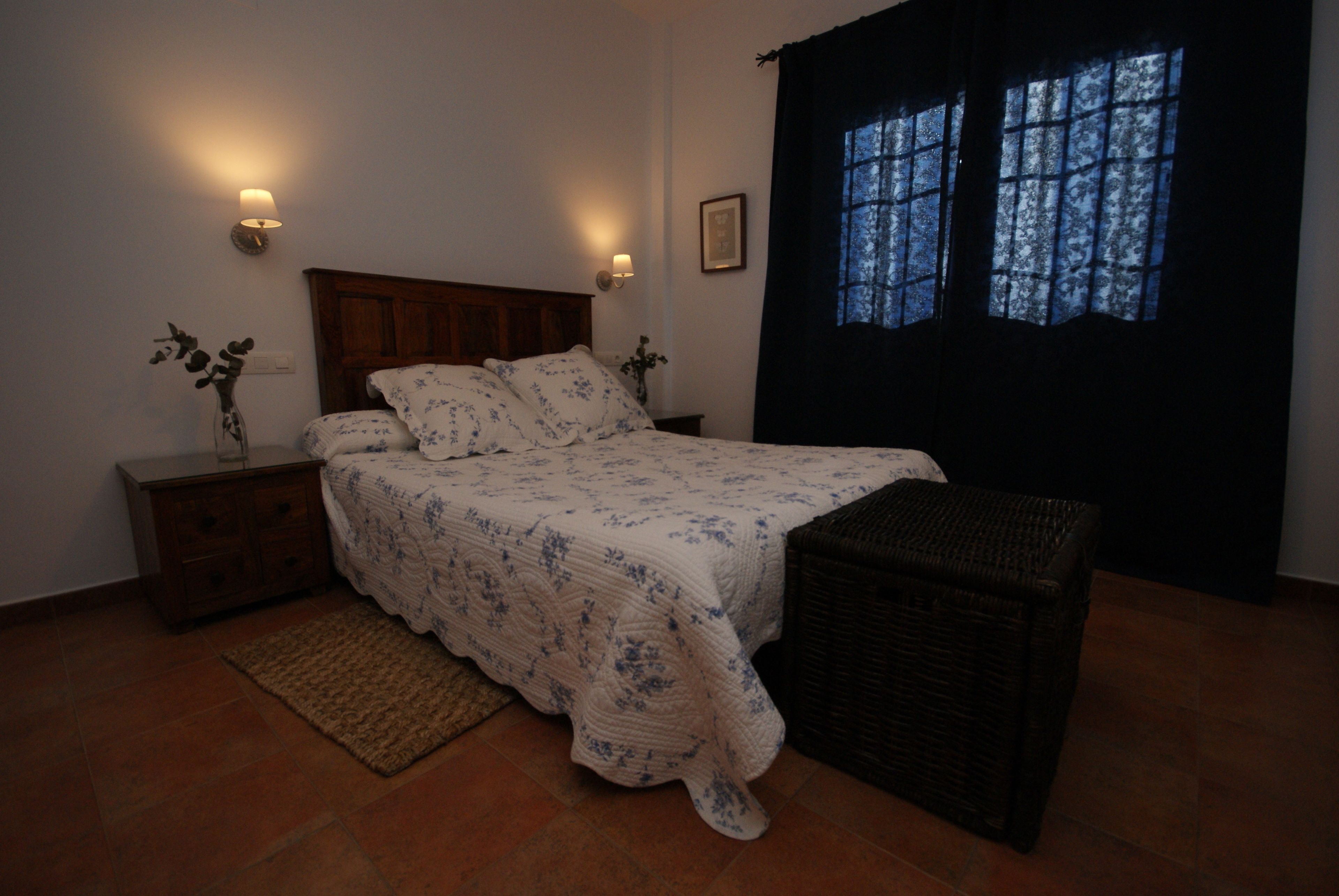 Un dormitorio con cama, mesita de noche, lámpara y ventana con cortinas.