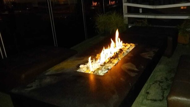 Un foyer extérieur rectangulaire, doté d’une flamme centrale et entouré d’une bordure en pierre, le tout se détachant sur un fond sombre.