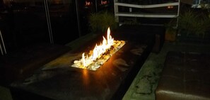 Una estructura de fuego exterior rectangular con llama central, rodeada por un borde de piedra, sobre un fondo oscuro.