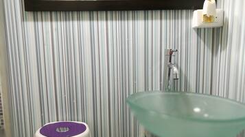 Un baño con pared a rayas, un lavabo de vidrio, un dispensador de jabón blanco y un cubo de basura morado.