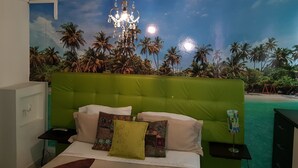 Une chambre avec une tête de lit verte, un lit agrémenté de plusieurs oreillers, une table de chevet et une fresque murale représentant une scène de plage.