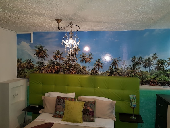 Un dormitorio con cabecera verde, cama con muchos almohadones, mesita de noche y un mural en la pared que representa una playa.