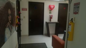 Un pasillo con dos puertas, una mesa pequeña con un florero, una decoración roja con forma de corazón y un extintor.