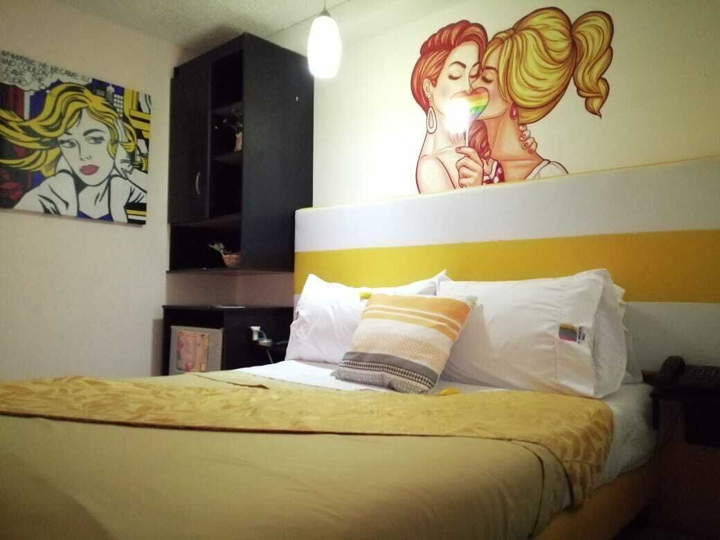 Una habitación de hotel con cama, cabecero, una pared decorada con obras de arte y un estante con objetos.