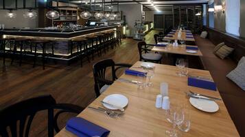 Un restaurante con una larga mesa de comedor dispuesta con platos, cubiertos y vasos, una zona de bar con taburetes, y un área de estar con sofás.