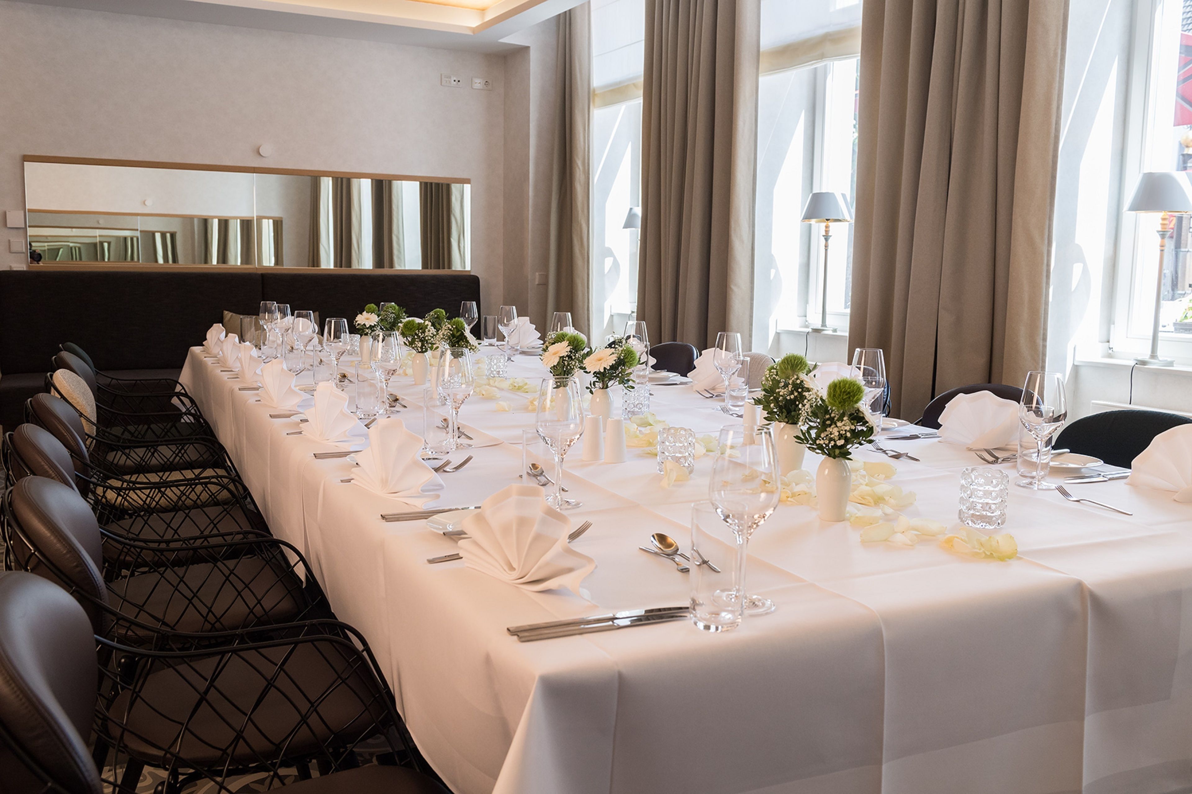 Una larga mesa de comedor dispuesta con manteles blancos, cristalería y centros de mesa florales en una habitación de hotel moderna con ventanas grandes y cortinas.
