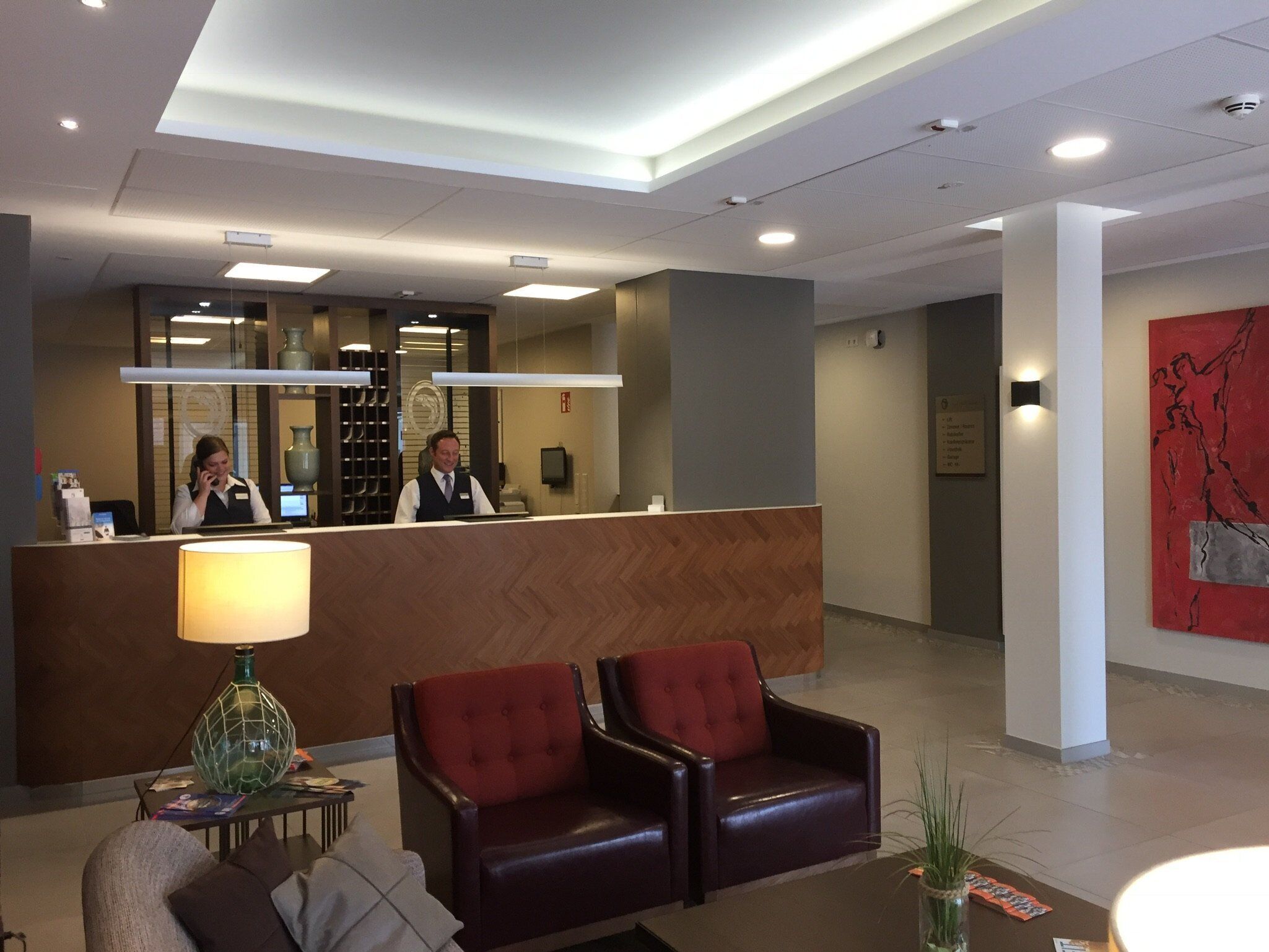 Et hotellets receptionområde med en receptionsdisk, to læderarmstole, en lampe og et stort abstrakt maleri på væggen.