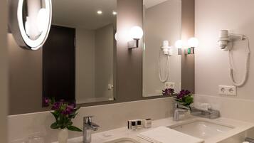 A modern bathroom with a large mirror, two sinks, a hairdryer, and a vase with flowers.