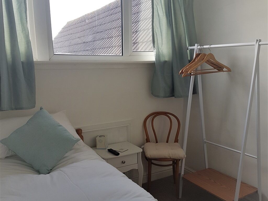 Un dormitorio con cama, mesita de noche, silla, perchero de madera y ventana con cortinas.