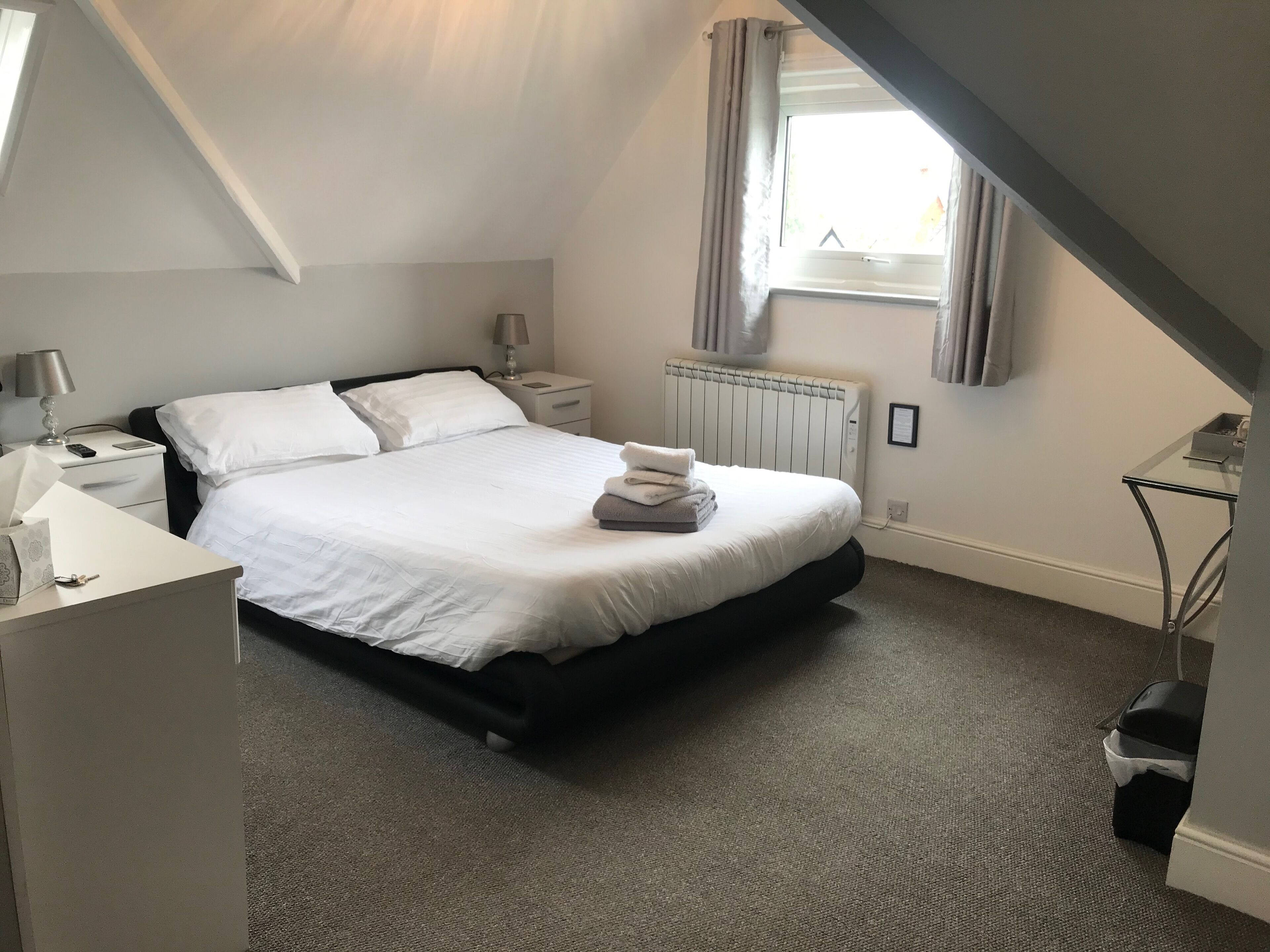 A neatly arranged bedroom with a bed, bedside tables, a lamp, a radiator, and a window with curtains.