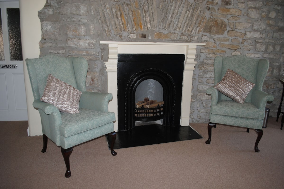 Two armchairs with patterned cushions, a fireplace with a stone wall backdrop, and a 'LAVATORY' sign on the left.