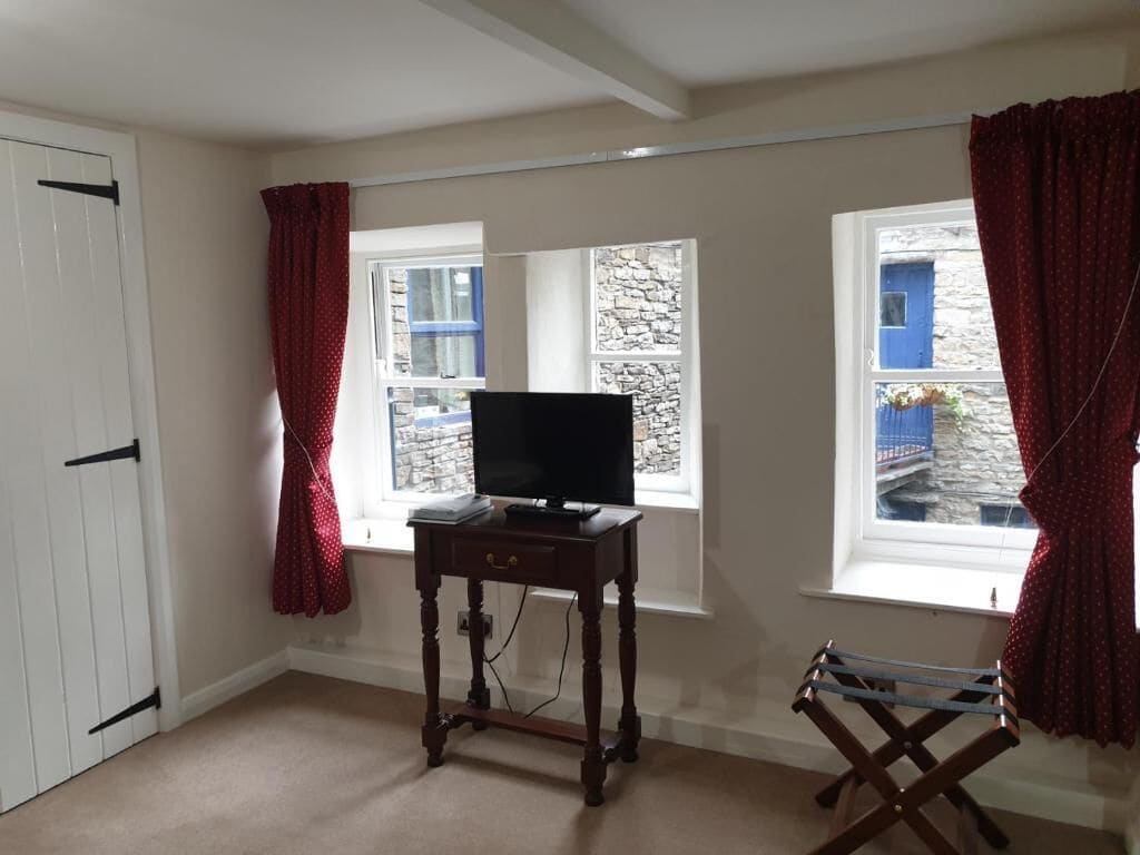 A room with a television on a wooden table, two windows with curtains, and a door.