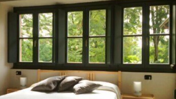 A bedroom with a bed, pillows, a nightstand, and a window with a view of greenery. - Ostello Costa Alta (Monza)