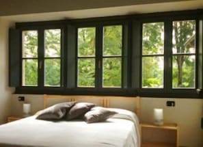 Un dormitorio con cama, almohadas, mesita de noche y ventana con vistas al verdor.