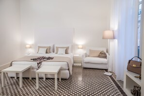 A hotel room with a large bed, two bedside tables, a sofa, a lamp, a handbag, and a shoe rack.