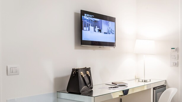 A modern hotel room with a glass desk, a white chair, a TV mounted on the wall, and a lamp on the desk. - Home Boutique Luxury & Design (Reggio Calabria)