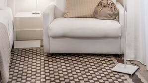 A hotel room with a white sofa, a bedside table with a lamp, a patterned rug, and a small bedside table with a remote control. - Home Boutique Luxury & Design (Reggio Calabria)