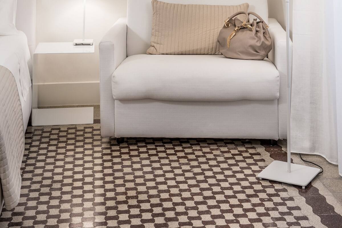 A hotel room with a white sofa, a bedside table with a lamp, a patterned rug, and a small bedside table with a remote control.