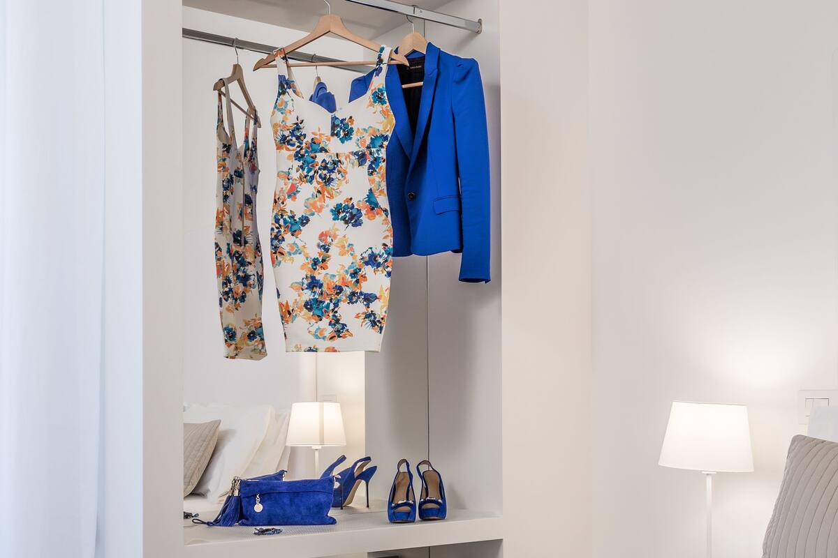 A hotel room with a bed, a bedside table with a lamp, a wardrobe with clothes, and a pair of shoes on the table.