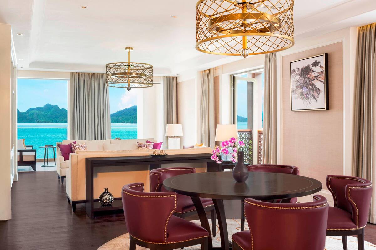 Un vaste espace de vie comprenant un canapé, une table de salle à manger ronde et des chaises recouvertes d’un tissu rouge. Une grande fenêtre offre une vue sur la mer et les montagnes.