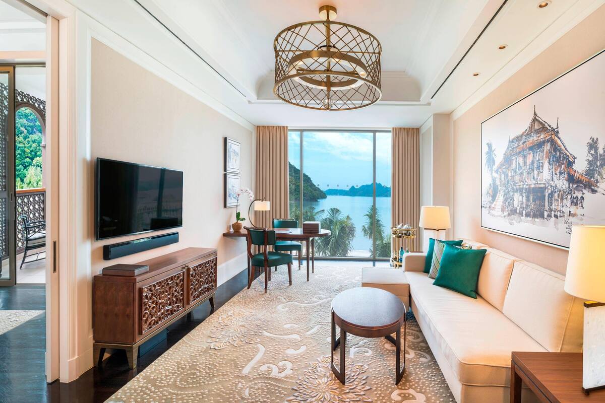 Une chambre d’hôtel dotée d’un grand téléviseur à écran plat, d’une console en bois, d’un canapé, d’un coin repas avec une table et des chaises, et d’une grande fenêtre offrant une vue sur un plan d’eau et la verdure.