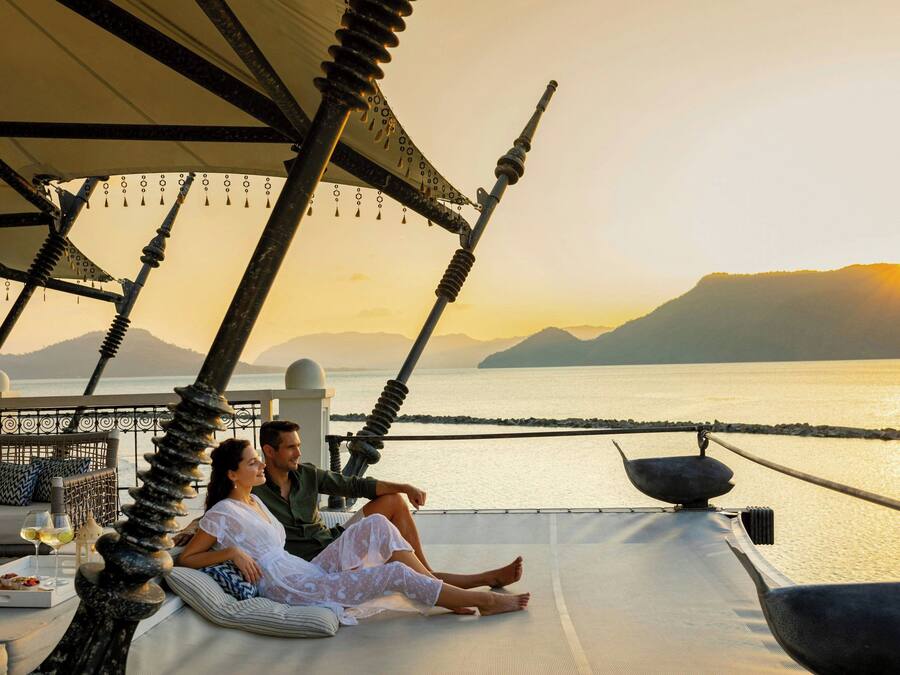 A couple relaxing on a sunlit deck with a view of the sea and mountains.