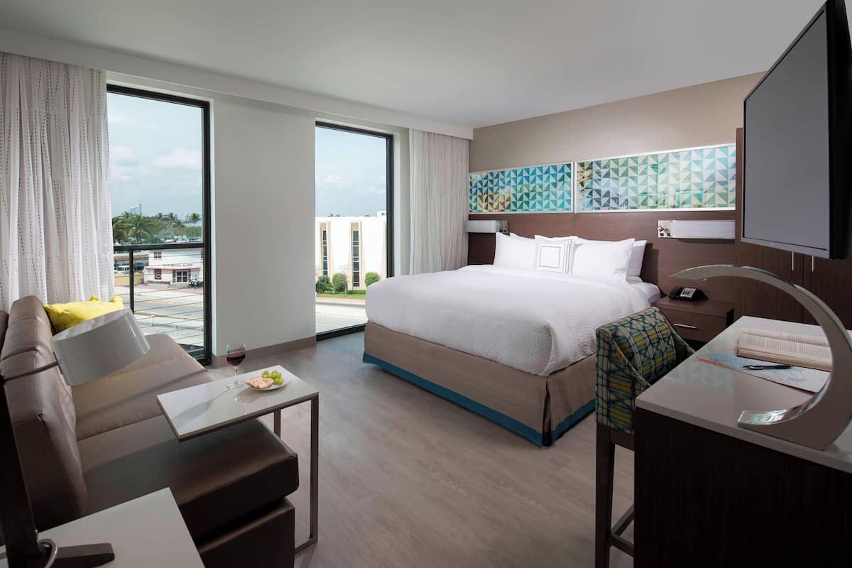 A modern hotel room with a large bed, a sofa, a desk, and a view of the city.