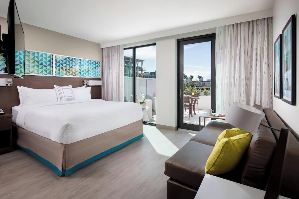 A modern hotel room with a large bed, a sofa, a balcony with a table and chairs, and a view of the city.