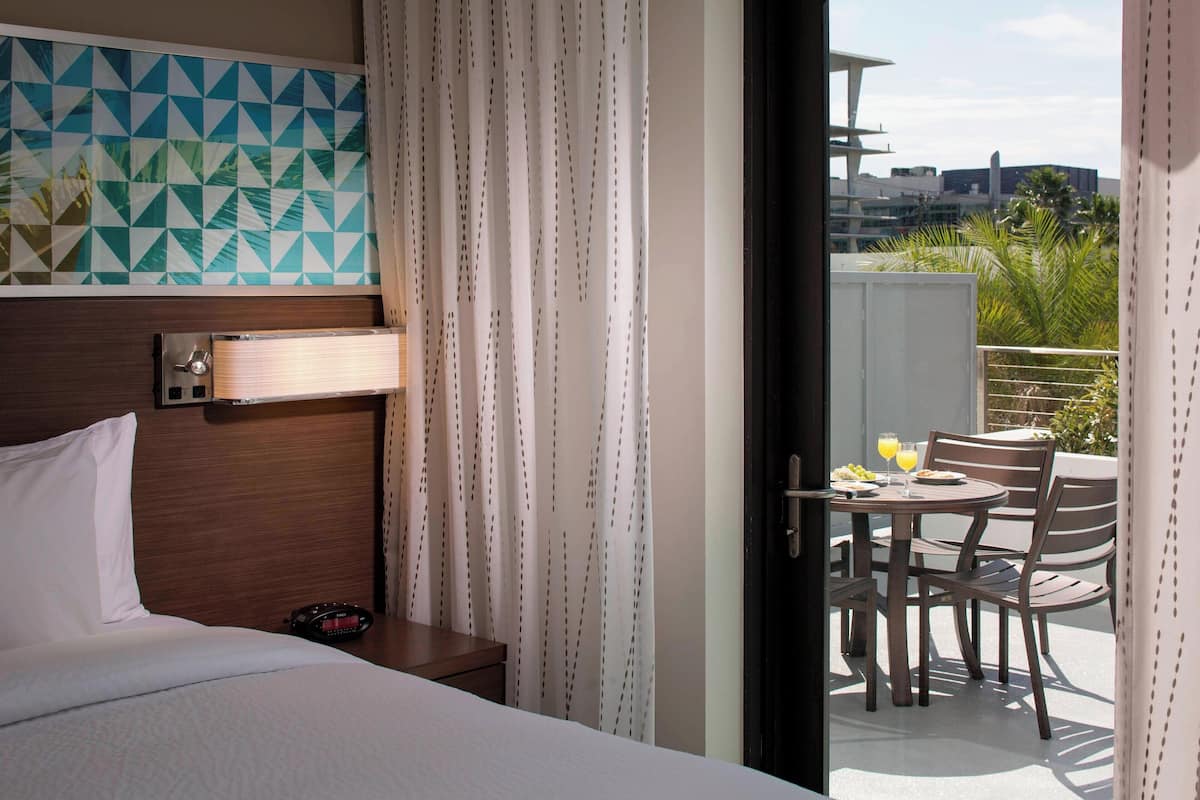 A hotel room with a bed, a bedside table, a wall-mounted alarm clock, and a view of a balcony with a table and chairs.