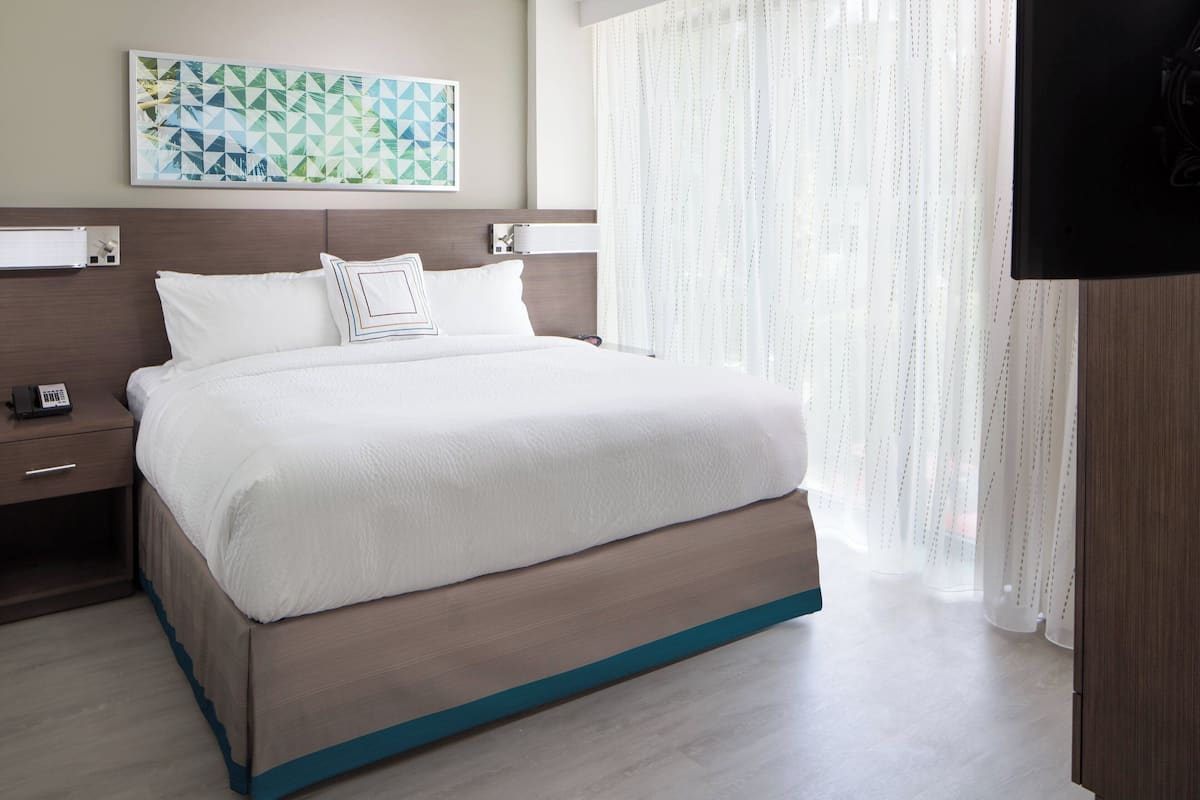 A neatly made bed with a wooden frame and white bedding, a bedside table with a phone, and a wall-mounted TV.