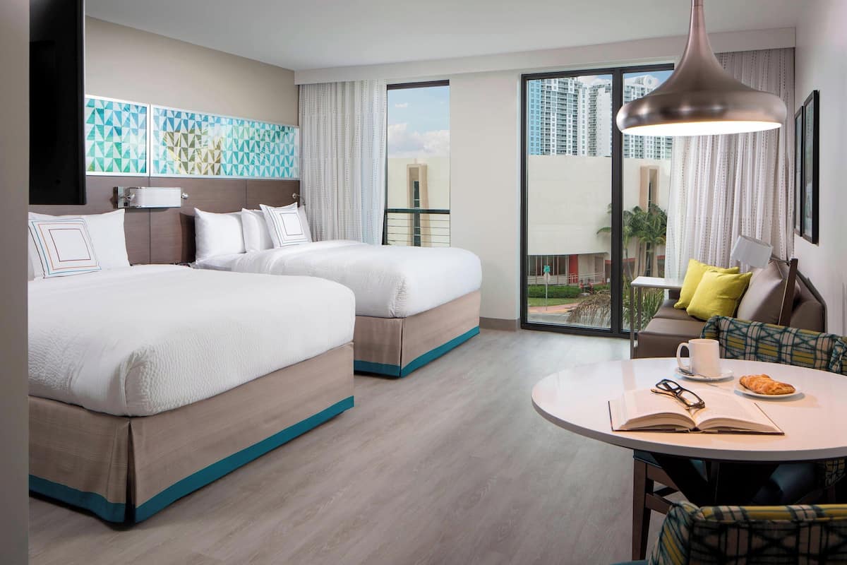 A hotel room with two beds, a dining table with a book and a cup, and a view of the city through large windows.