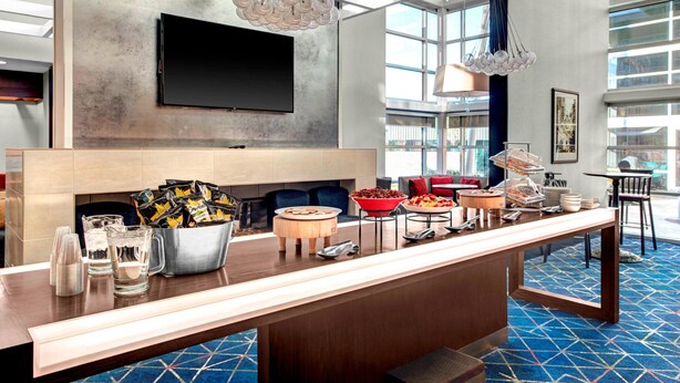 A modern hotel lobby with a long reception counter, a flat-screen TV mounted on the wall, and a buffet area with various food items. - Residence Inn by Marriott Philadelphia Airport (Philadelphia)