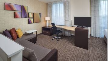 Habitación de hotel con sofá, escritorio, silla, televisor y obra de arte en la pared.