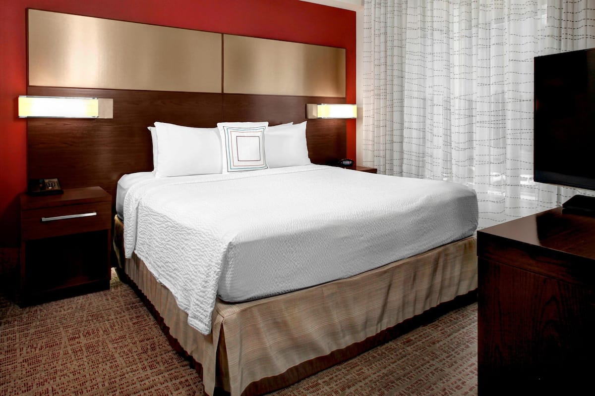 A hotel room with a large bed, wooden headboard, two bedside tables, a TV, and a window with sheer curtains.