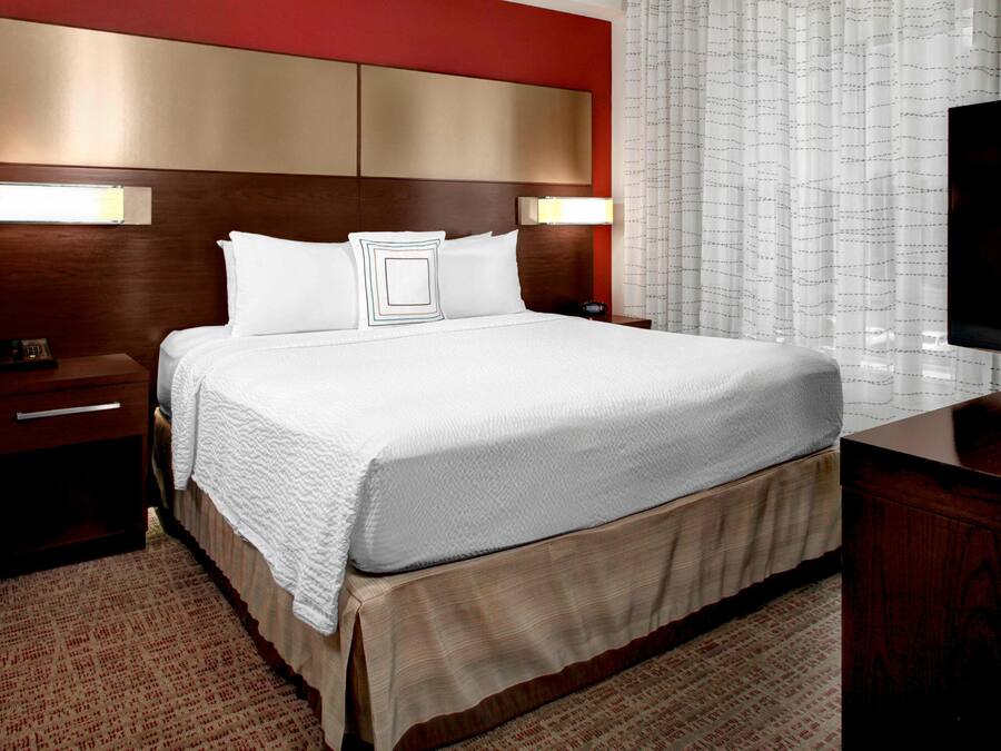 Una habitación de hotel con una cama grande, cabecera de madera, dos mesitas de noche, un televisor y una ventana con cortinas translúcidas.