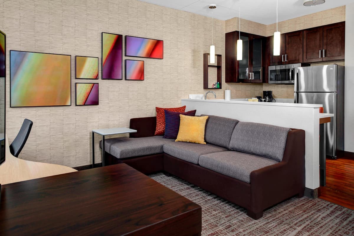 A modern kitchen with a built-in microwave, refrigerator, and a small island. There is a dining area with a wooden table and a sofa with colorful cushions.