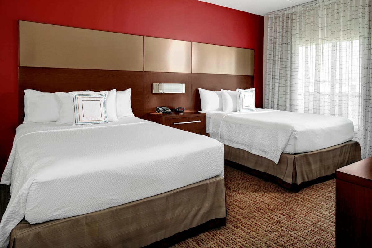 A hotel room with two beds, a wooden headboard, a nightstand with a phone, and a window with sheer curtains.