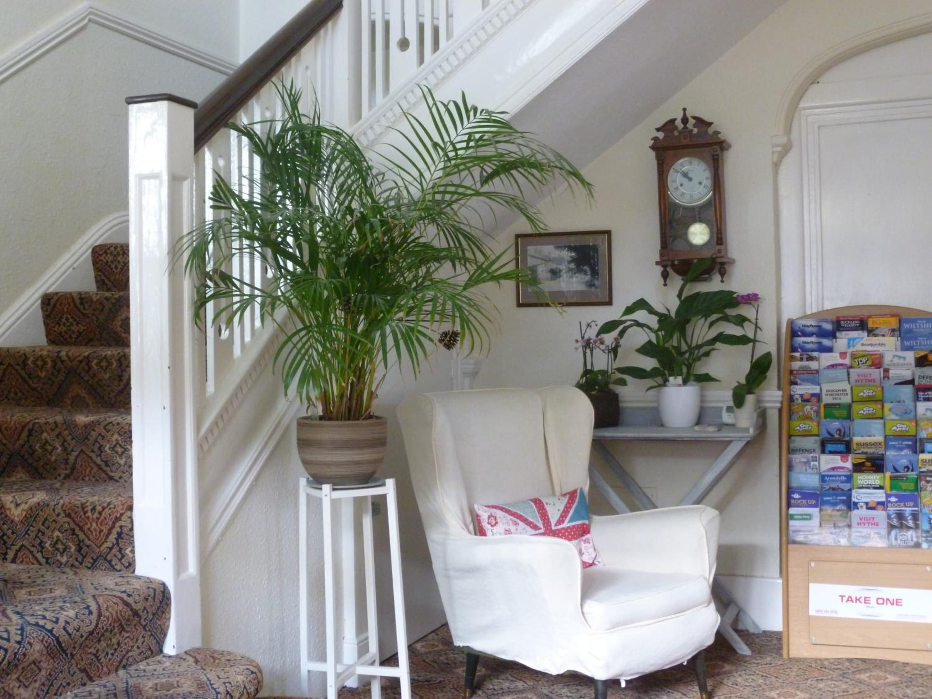 Un escalier recouvert d’une moquette à motifs, une chaise blanche avec un coussin, une plante en pot, une horloge et un panneau d’affichage avec des brochures.