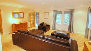 A spacious living room with a large sectional sofa, a small coffee table, and a fireplace. There is a wooden cabinet and a bookshelf in the corner. - BCC Lochness Cottages (Inverness)