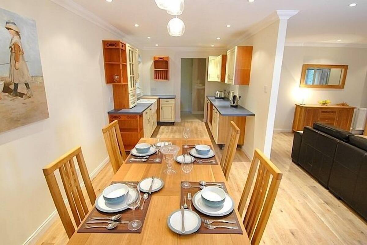 A modern kitchen with wooden cabinets, a dining table set for six, and a black sofa in the living area.