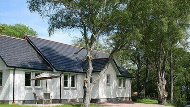 A single-story house with a dark roof, white walls, and a small porch with a chair. - BCC Lochness Cottages (Inverness)