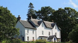 A white two-story house with a steeply pitched roof, surrounded by lush greenery and trees.