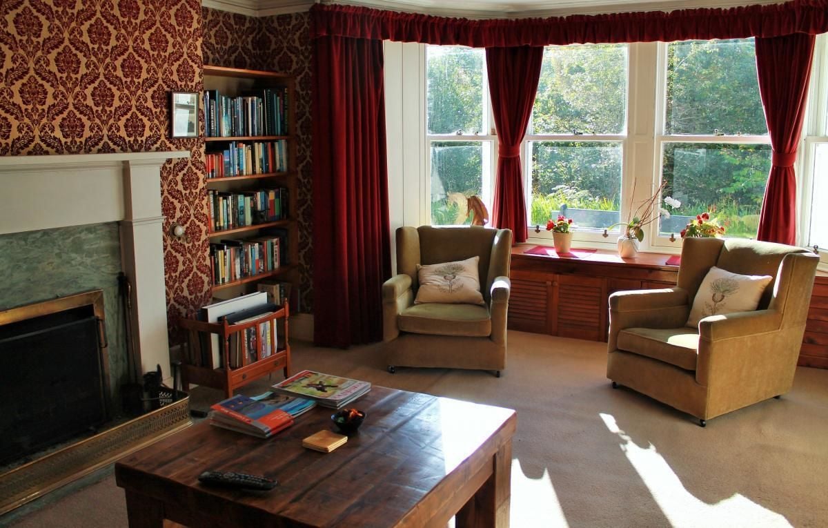 A cozy living room with a fireplace, a bookshelf, two armchairs, a coffee table, and large windows with curtains.