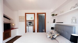 Habitación de hotel con cama, mesita de noche, lámpara y vista al área de la cocina a través de una puerta abierta.