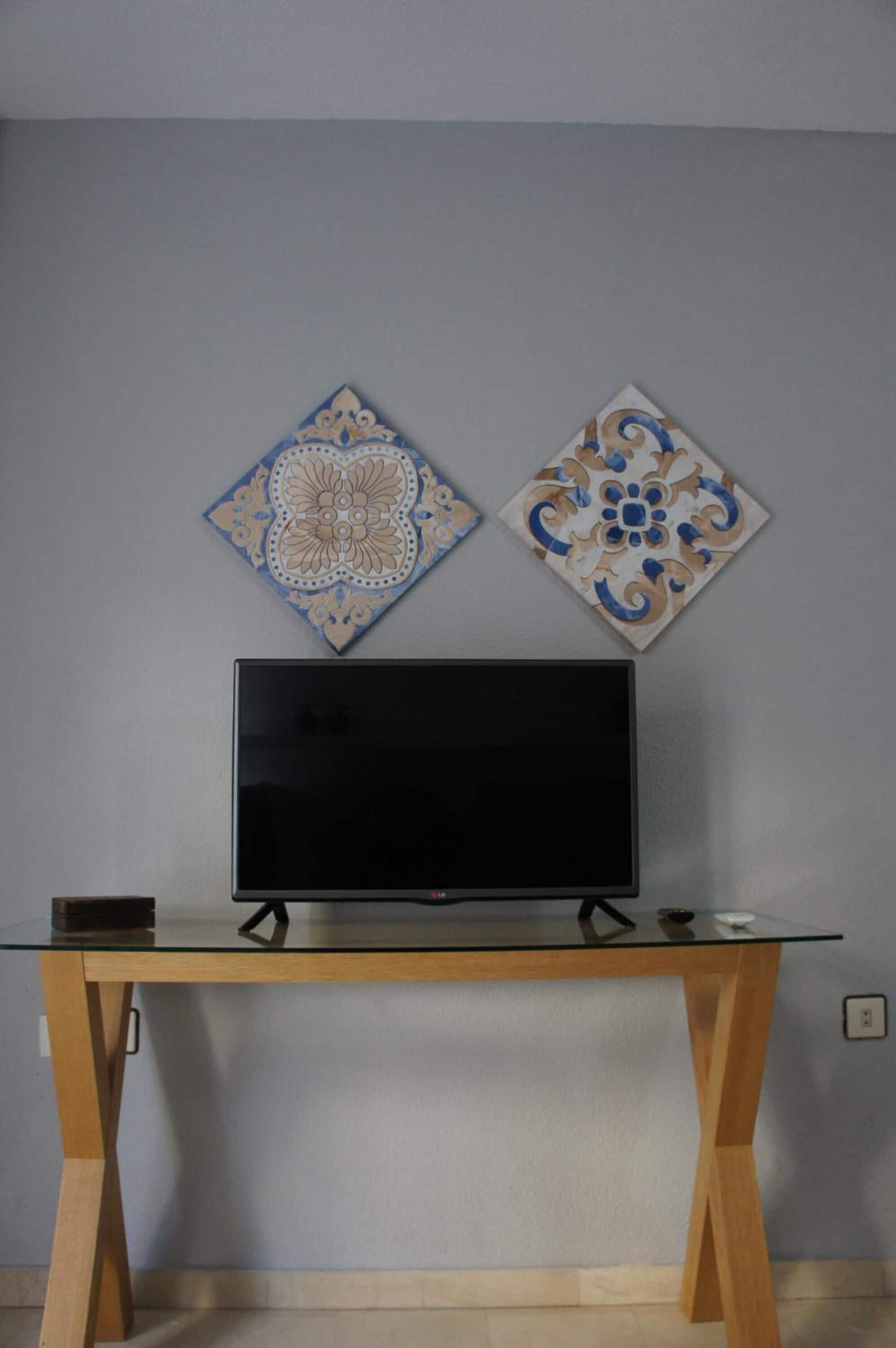 A flat-screen TV on a glass-top table with wooden legs, two decorative wall tiles above it, and a power outlet on the wall.
