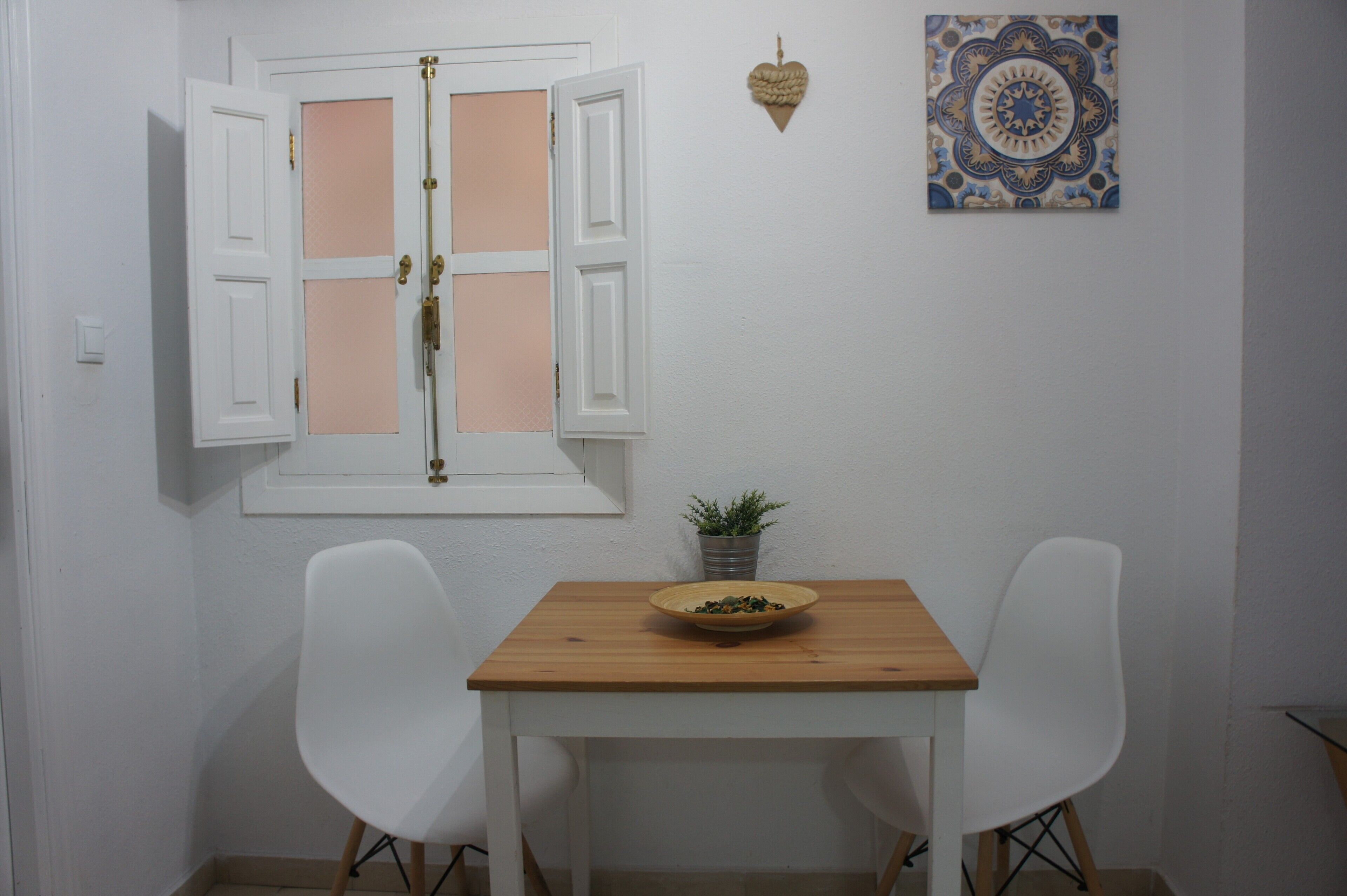 Una habitación pequeña con una mesa de madera, dos sillas blancas, una ventana con contraventanas abiertas, una planta en maceta y un azulejo decorativo en la pared.