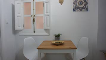 Una habitación pequeña con una mesa de madera, dos sillas blancas, una ventana con contraventanas abiertas, una planta en maceta y un azulejo decorativo en la pared.