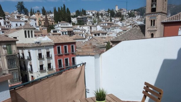 Una terraza en la azotea con una mesa y sillas de madera, una planta en maceta y vistas a un pueblo con edificios y una torre.