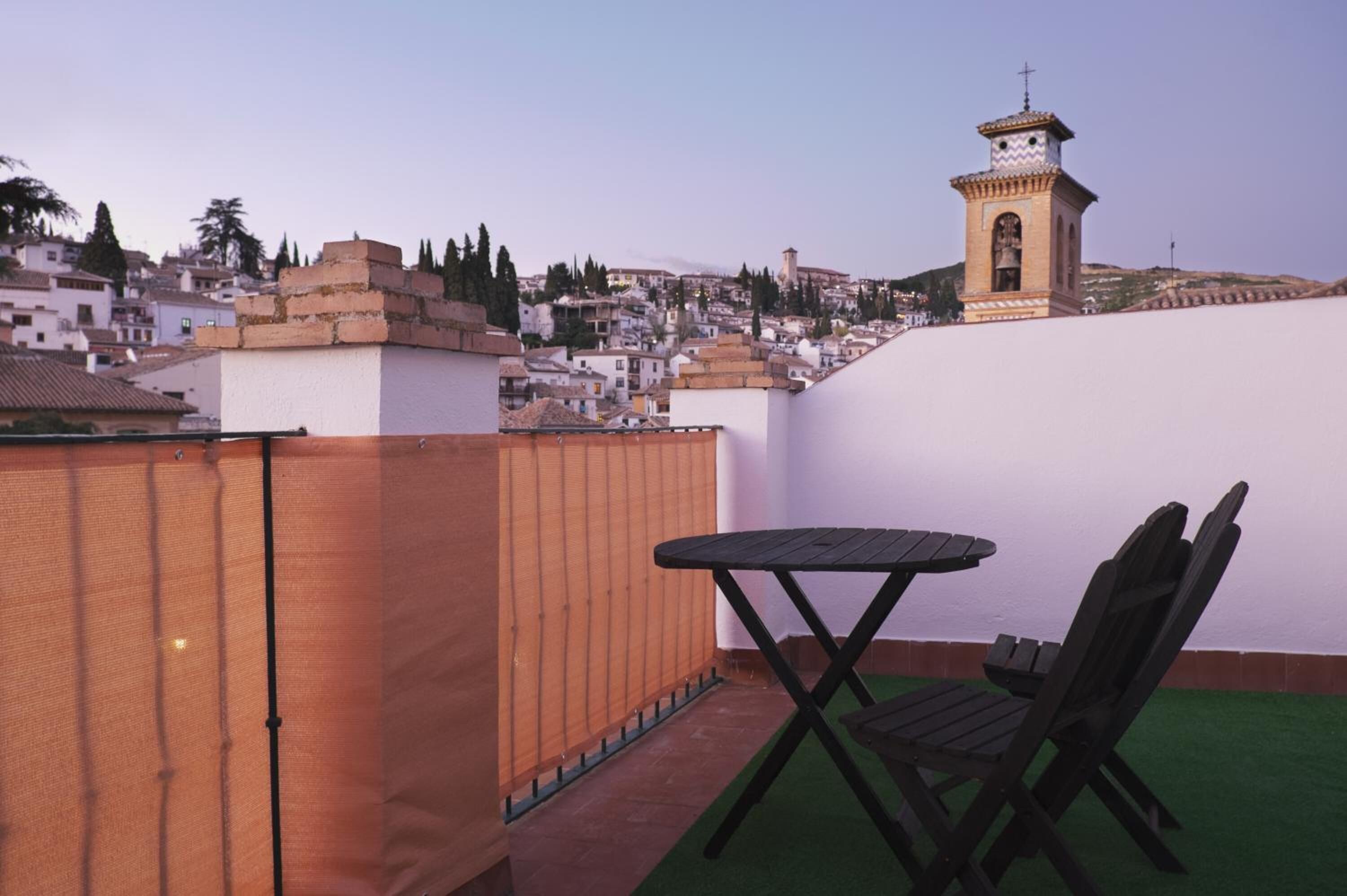 Terraza en la azotea con mesa y sillas, con vistas a un paisaje urbano donde destaca una torre.