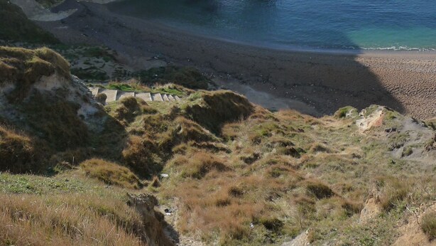A coastal landscape with a prominent rock formation arching over the sea, a sandy beach, and grassy terrain in the foreground. - The Esplanade (Weymouth)